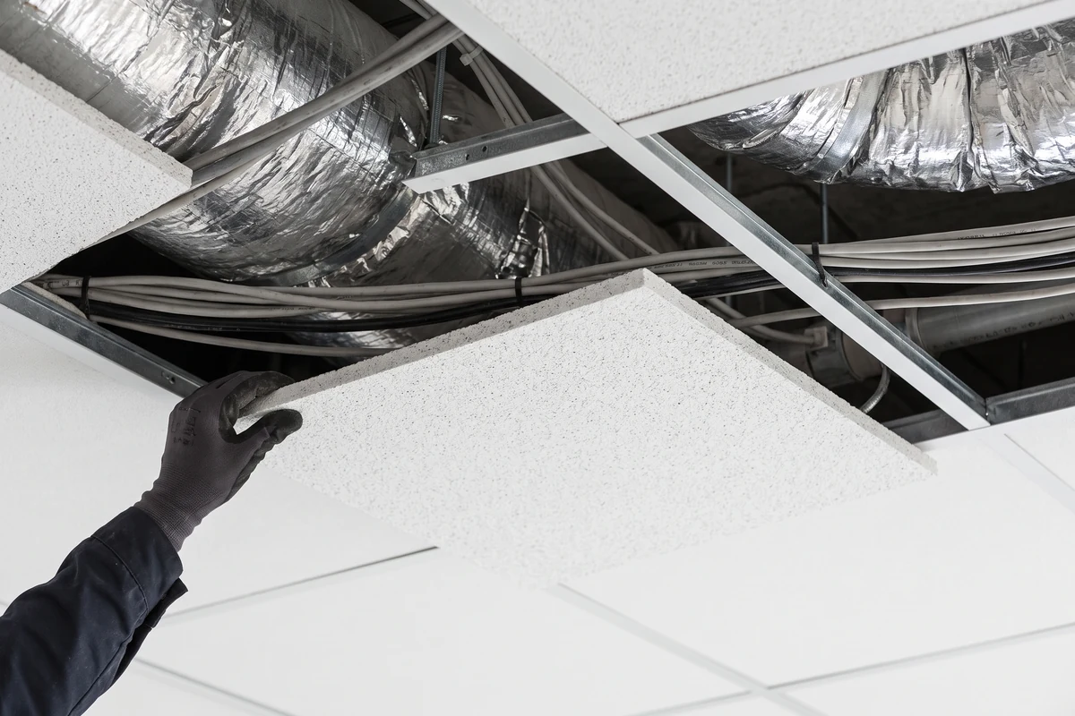 Hand die een plafondplaat van een systeemplafond optilt, met leidingen erboven